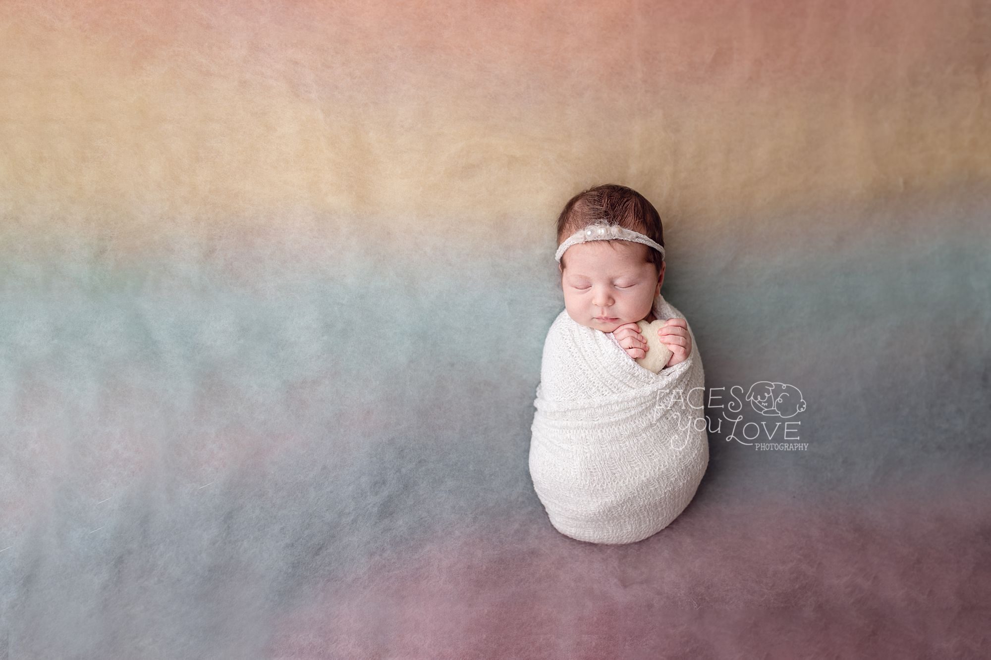 Baby girl on rainbow backdrop holding tiny heart by Kansas City newborn photographer