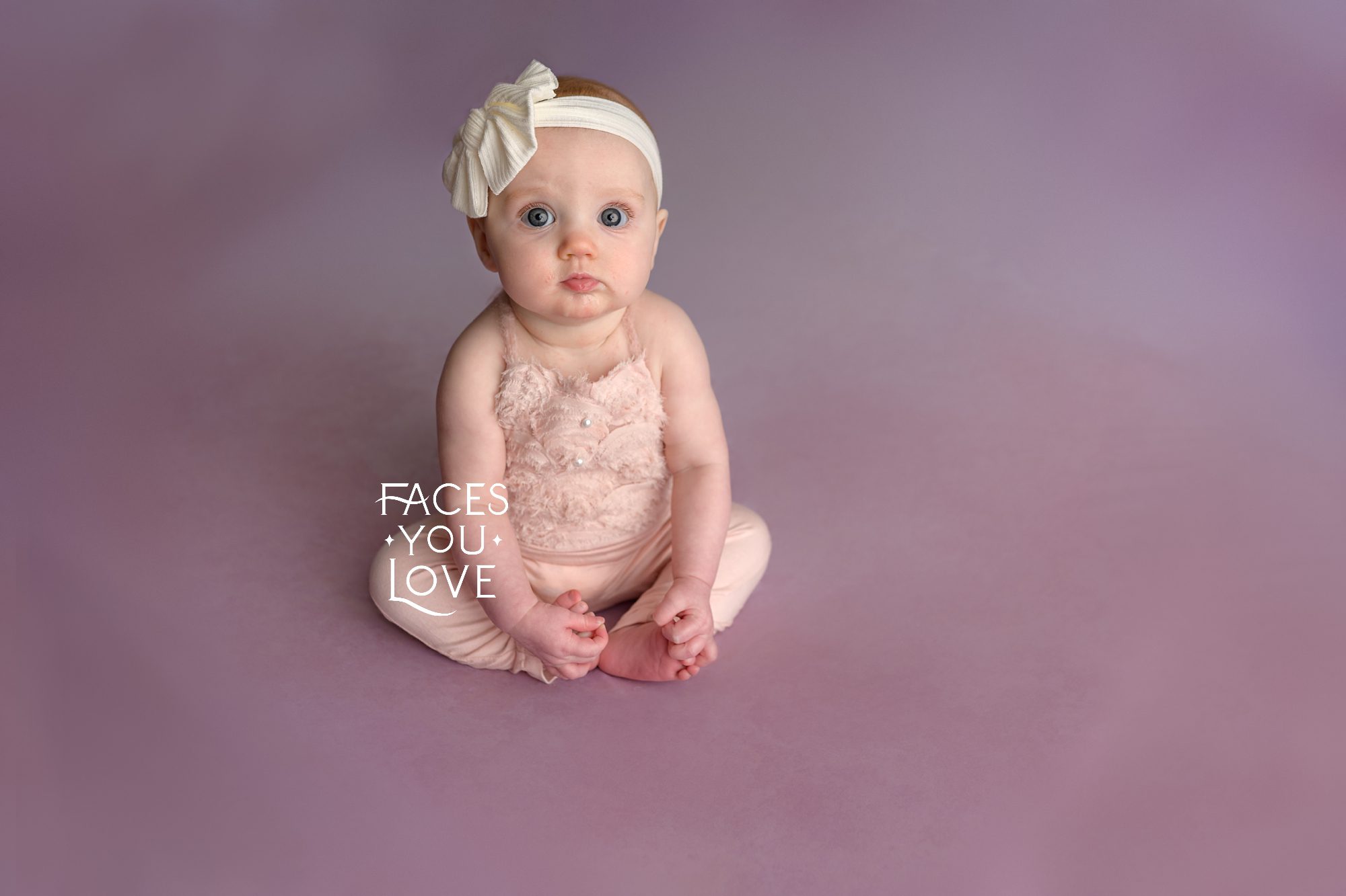 Six month old baby girl, sitting and holding her toes, on a pink backdrop. She's wearing a headband and has a serious expression. Photographed in Kansas City by Faces You Love Photography.