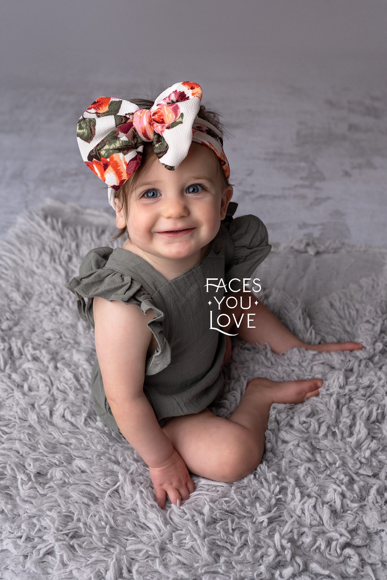 Baby girl sitting on gray fur, wearing a sage green romper and a big floral hair bow. Photographed by Helen Ransom of Faces You Love in Overland Park.