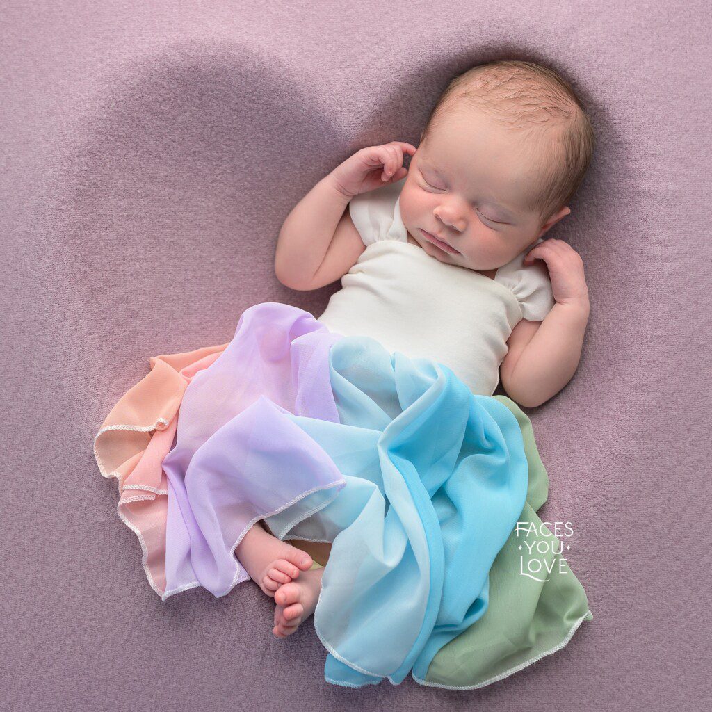 Newborn girl laying within a purple heart shape, and wearing a rainbow dress. Photographed in Overland Park by Faces You Love Photography.