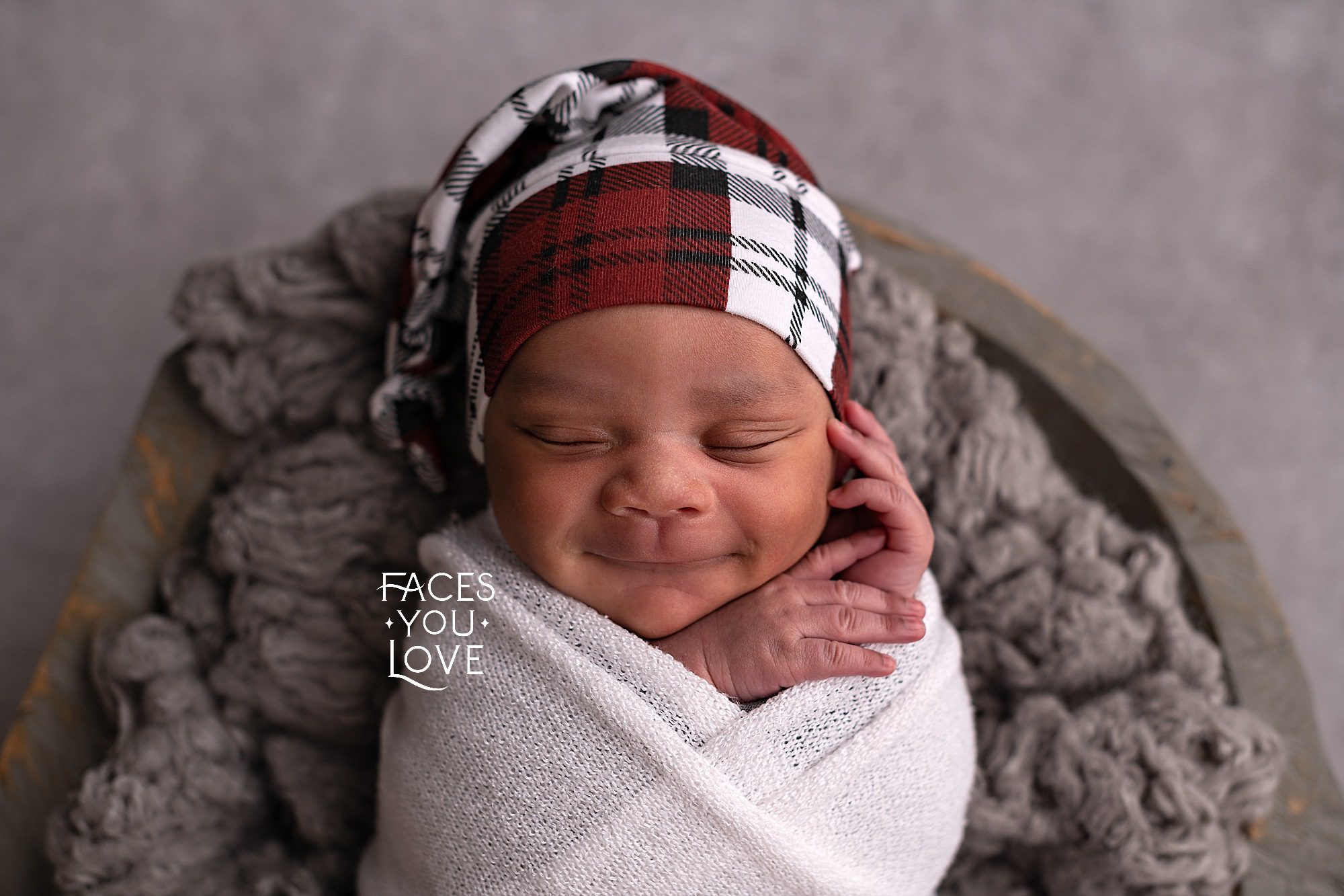 Smiling newborn baby, wearing a red, white, and black stocking cap, wrapped in a white wrap, and laying on gray fluff in a wooden gray bowl. Photographed by Helen Ransom of Faces You Love Photography.