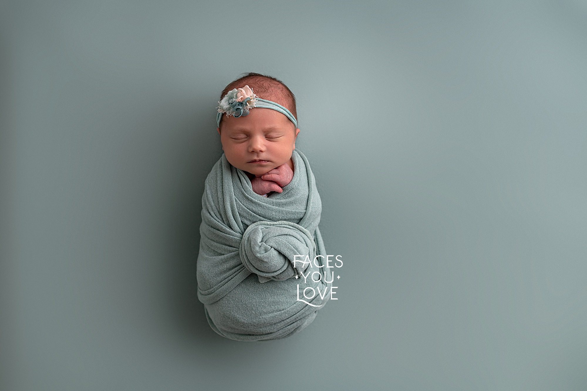 Newborn baby girl, on a tropical blue backdrop and wrapped in the same. The wrap is twisted into a knot at the front of the baby, and her hands are under her chin. Photographed in Kansas City.