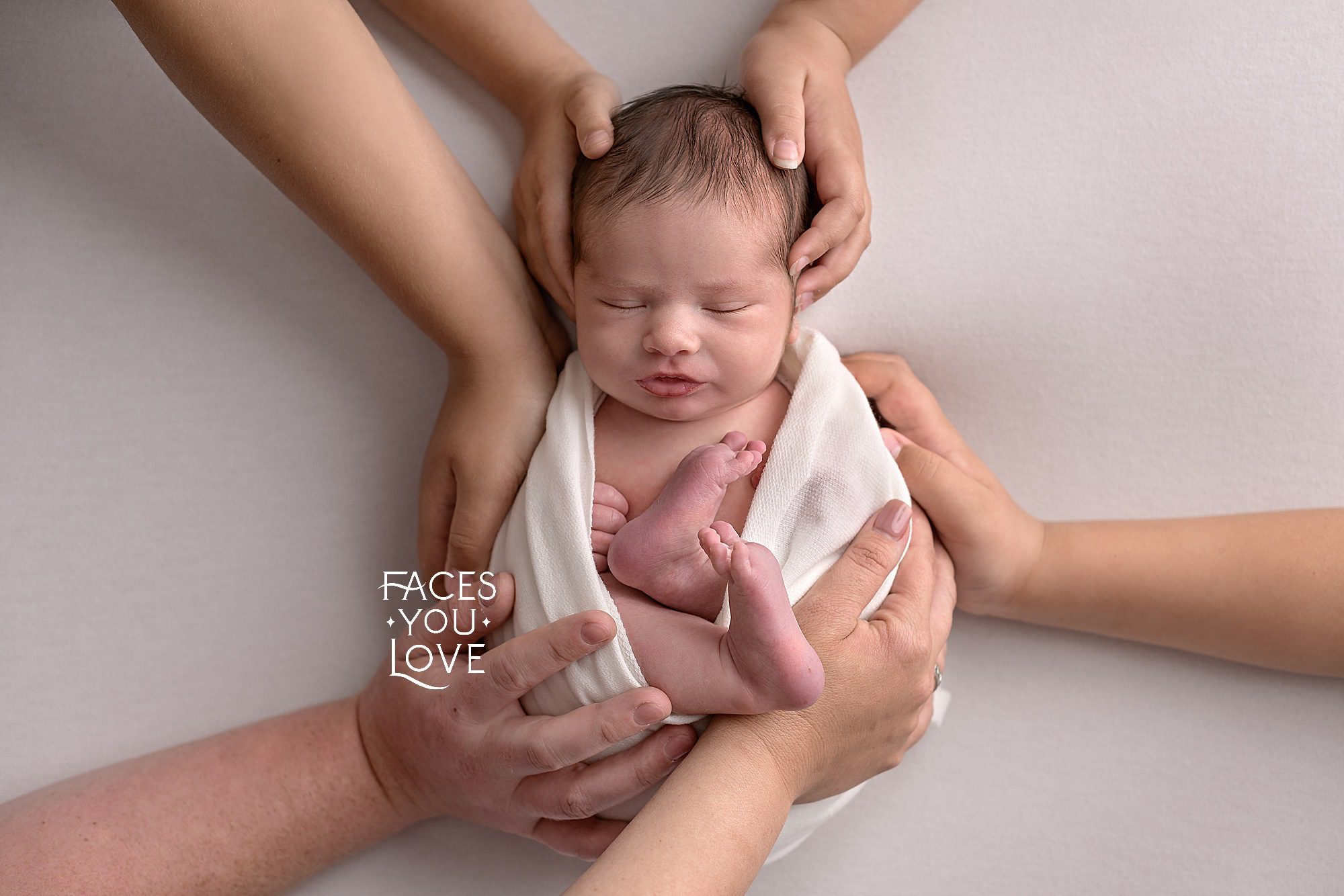 Creative pose of a newborn baby born into a large family. He's laying on white, and wrapped in white. He has 6 different hands laying on him, one from each of his family members. Photographed by Helen Ransom of Faces You Love in Kansas City.