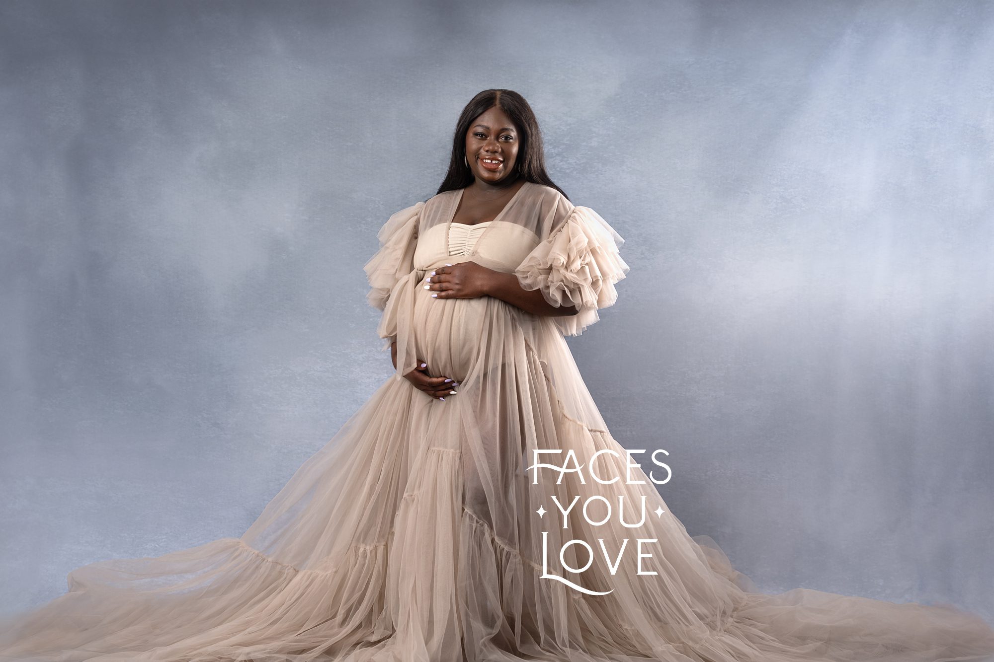 African American woman expecting a baby at her maternity photoshoot. She's wearing a frilly, sand colored robe and body suit, while standing on a light blue backdrop. Photographed in Overland Park by photographer Helen Ransom.