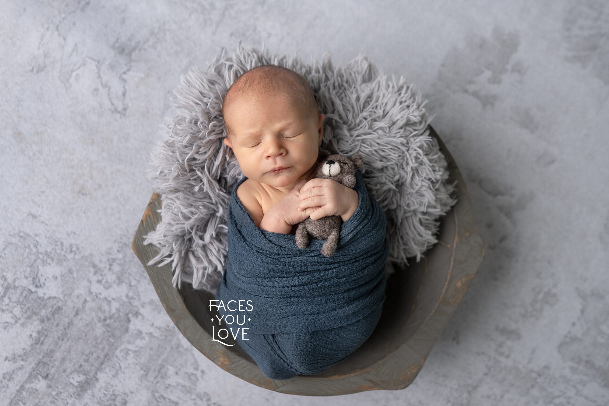 Newborn boy, wrapped in blue and laying in a gray fur lined wooden bowl. He's been posed to carefully hold a tiny wool teddy bear. Photographed by Helen Ransom of Faces You Love Photography in Overland Park, KS.