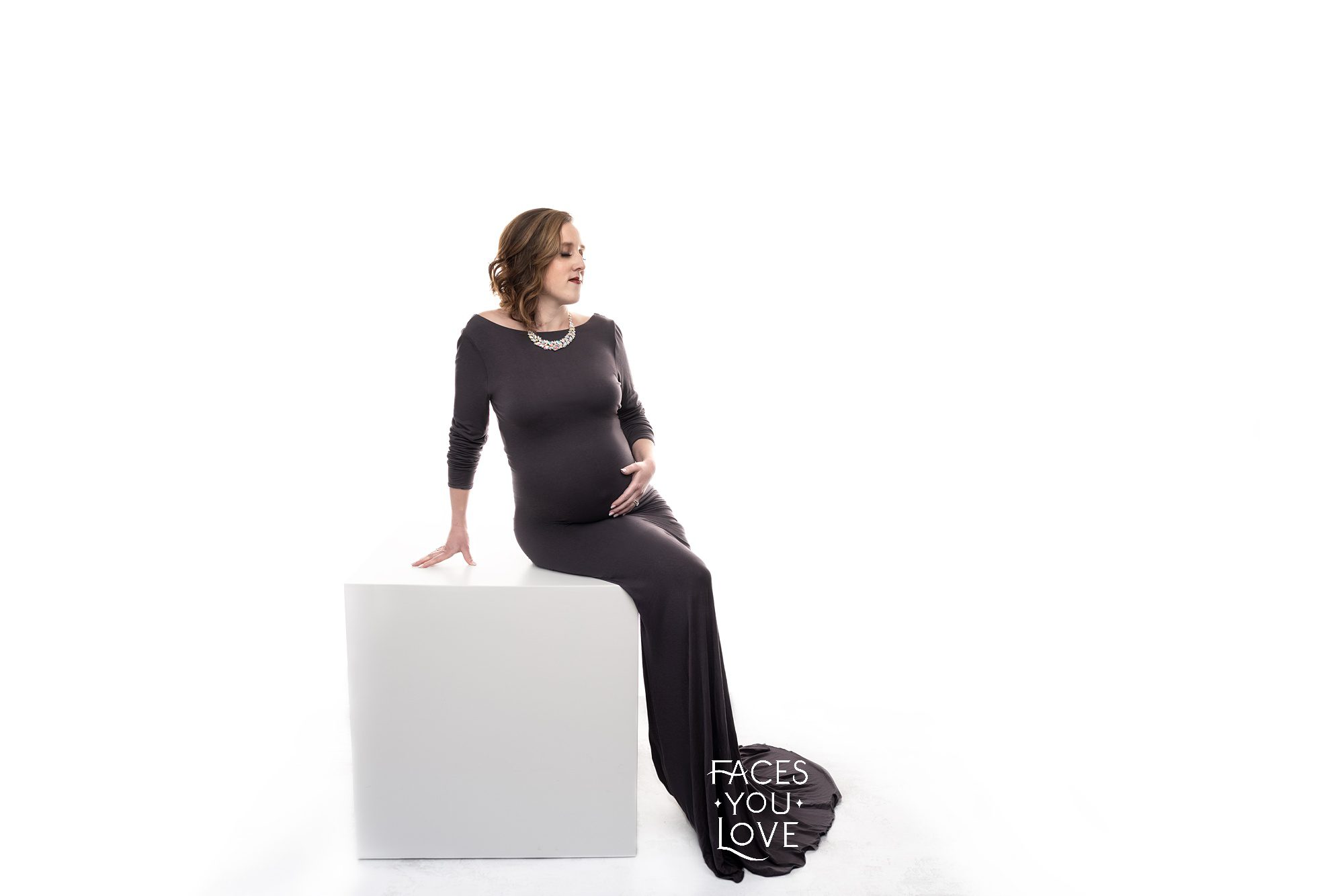 Glam maternity session photographed by Helen Ransom of Faces You Love Photography. Mom is in an elegant, long dark gray dress and wearing a statement necklace. She's on a white posing box, against a high-key white background.