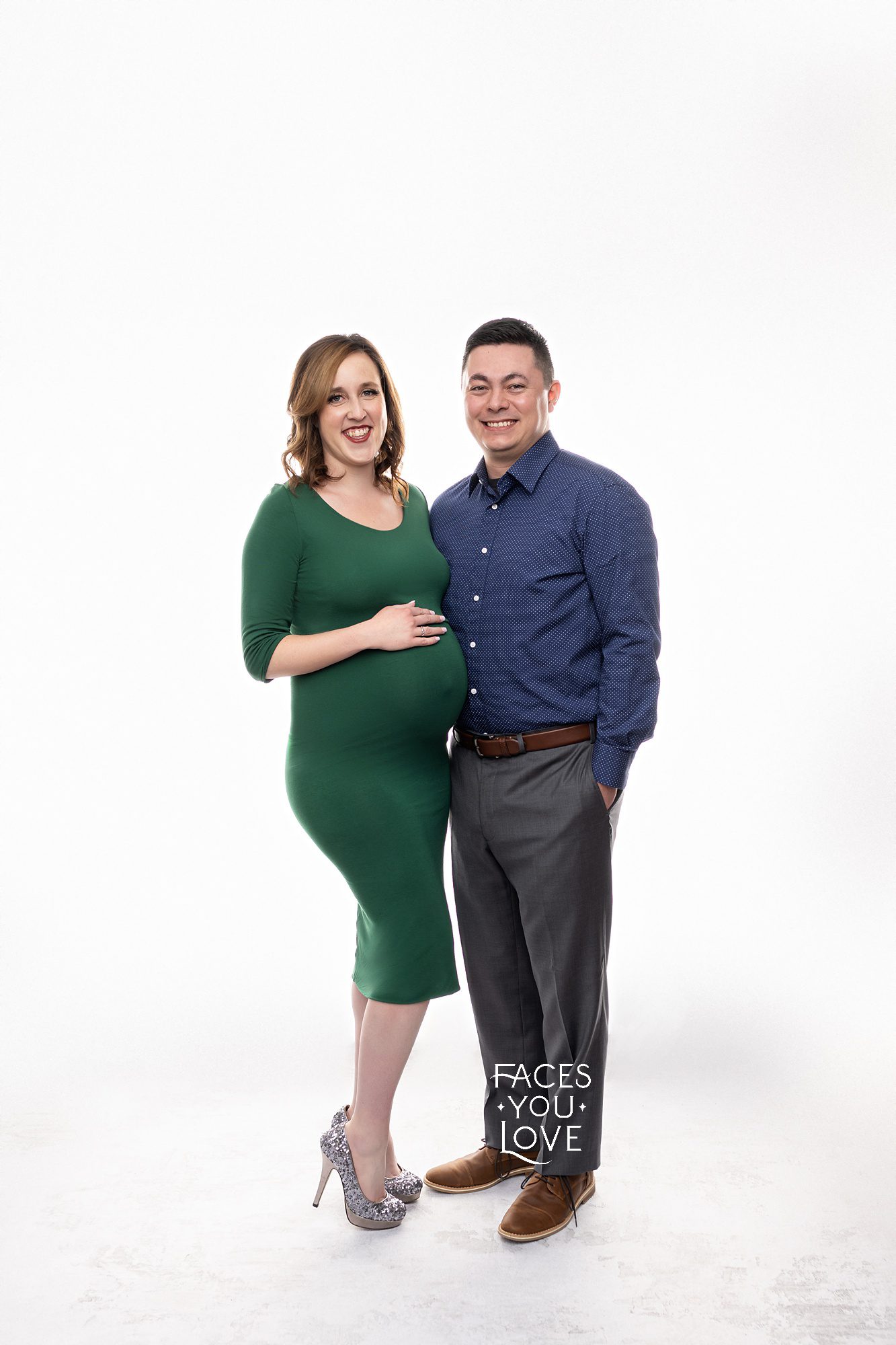 Maternity couple pose on high-key lighting, at Faces You Love studio in Kansas City. Pregnant mom is wearing an emerald green, cocktail length dress and dad is posed next to her in slacks and a button down shirt.