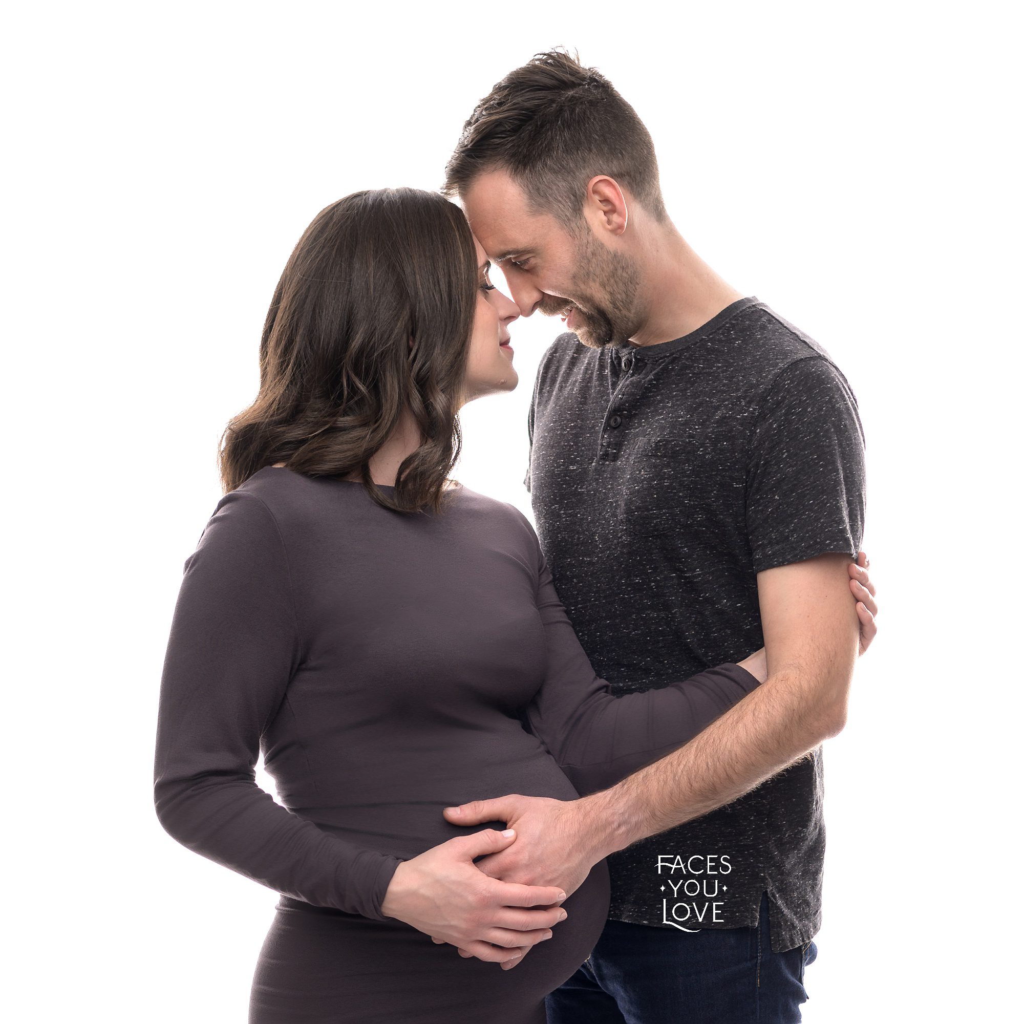 Studio maternity portrait of a couple. They are facing each other, foreheads and noses touching, and their arms are entwined around mom's belly. Photographed on a high-key white background, with both of them wearing dark gray. Photo made at the Faces You Love Photography studio in Overland Park.