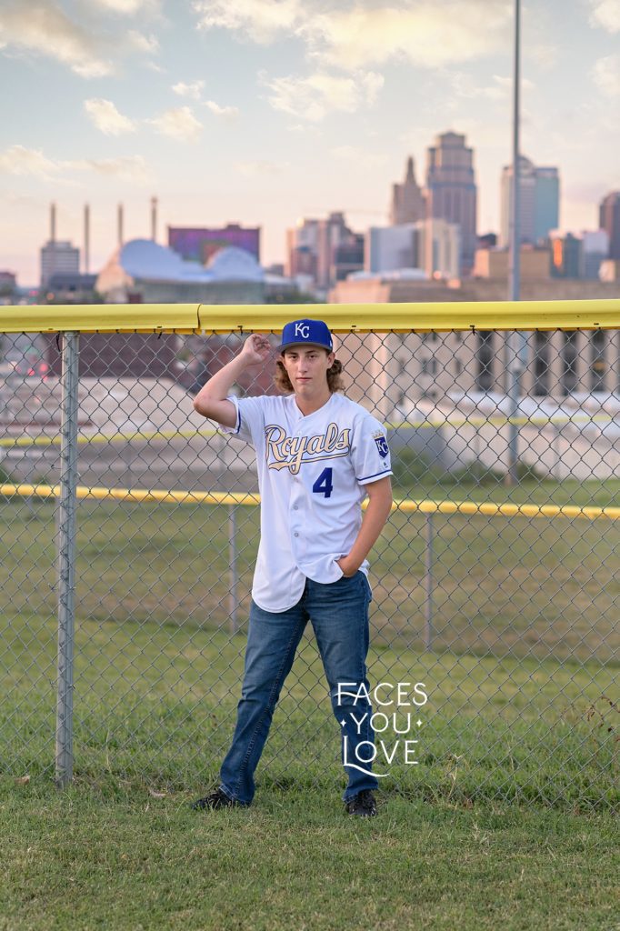 Senior boy, wearing a Kansas City Royals jersey and hat, at a baseball field overlooking the Kansas City skyline. Senior photo session photographed by Faces You Love Photography.
