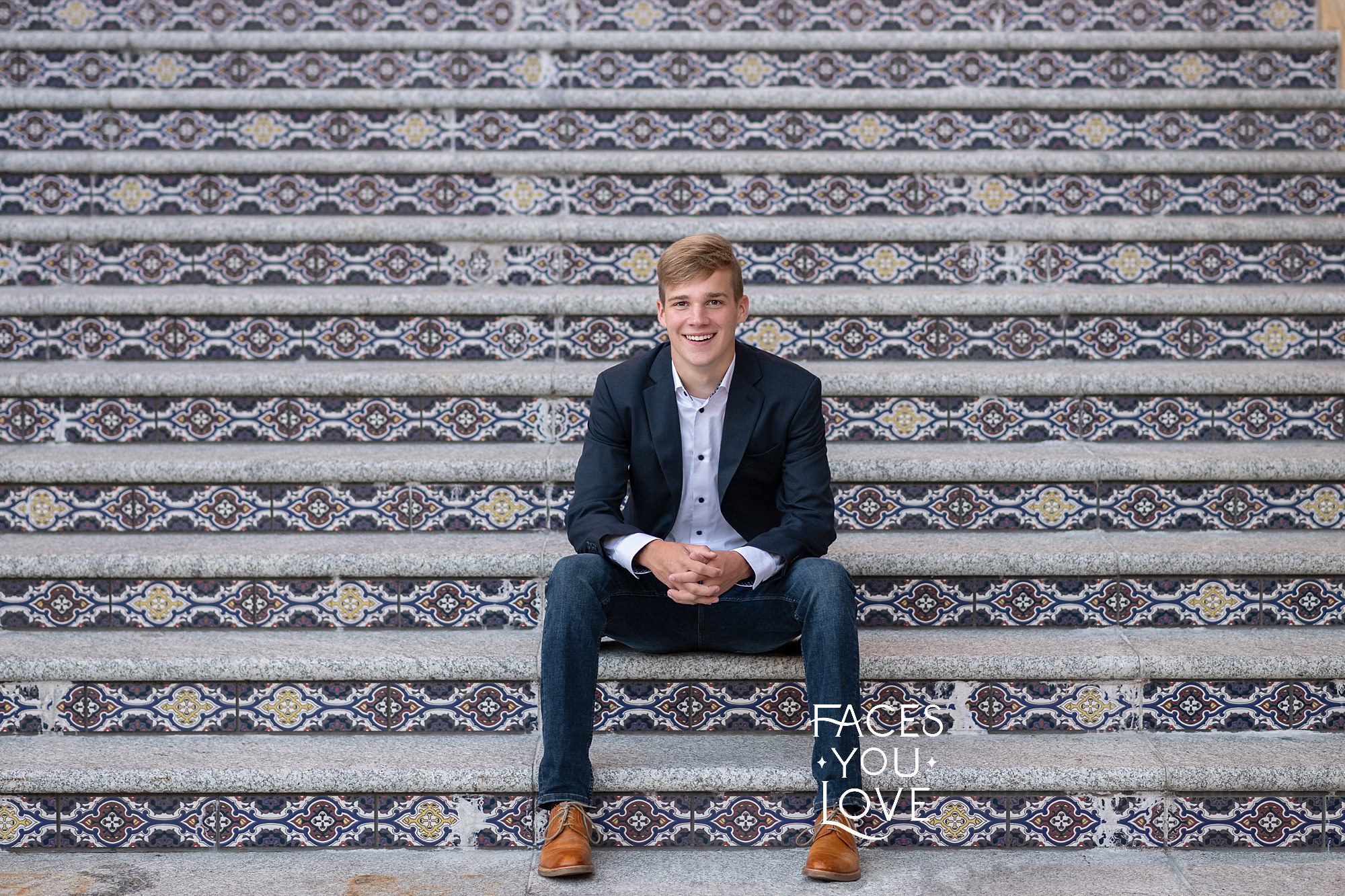 Senior boy picture, photographed by Faces You Love an Overland Park photography studio. Boy is sitting on the tile steps at the Country Club Plaza, while wearing jeans, a sport coat, and brown dress shoes.