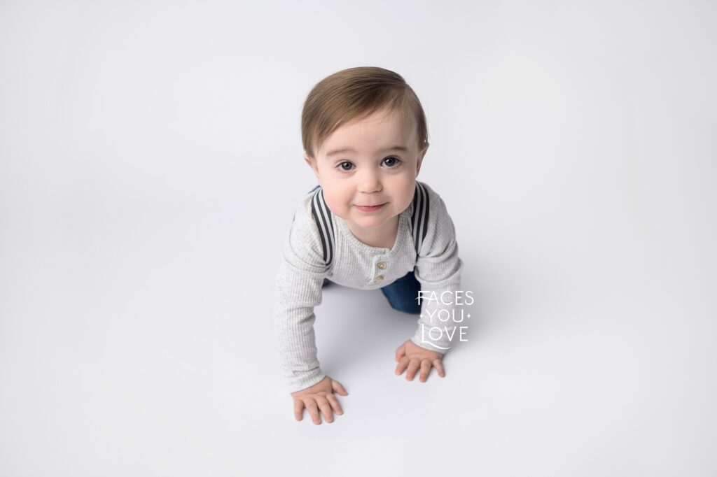 Baby crawling towards the camera, on a clean, white background. He's wearing a gray, waffle texture shirt and suspenders. Photographed by Helen Ransom of Faces You Love in Overland Park, KS.