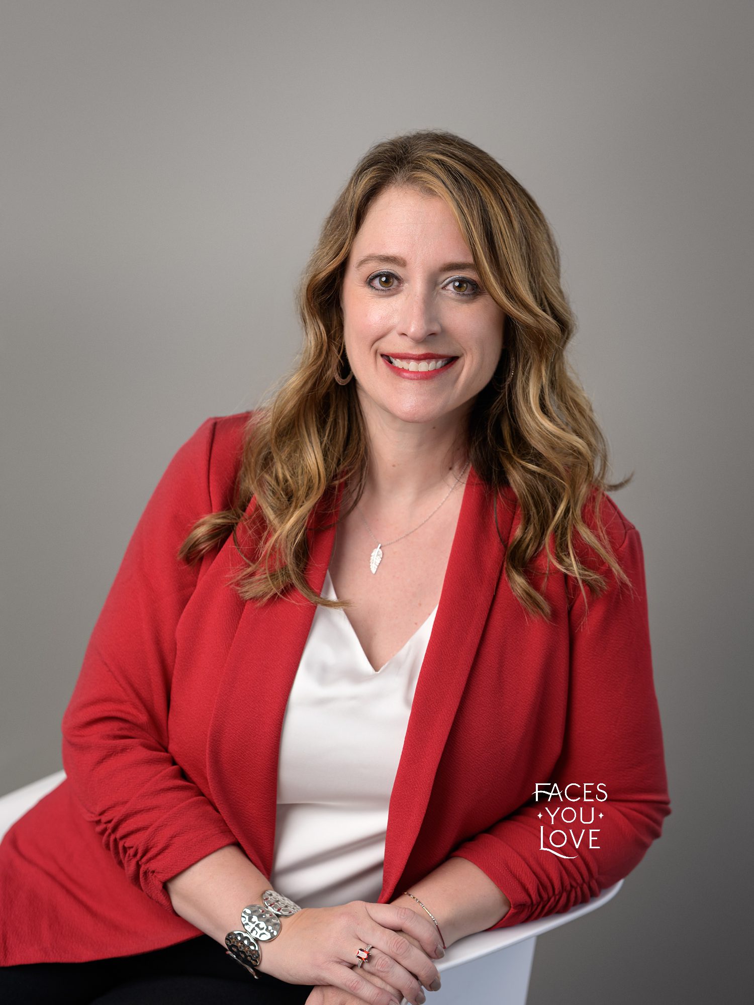 Professional female headshot, photographed on a gray background. She's sitting in a white chair and wearing a red blazer. Photographed at Kansas City headshot studio Faces You Love Photography.