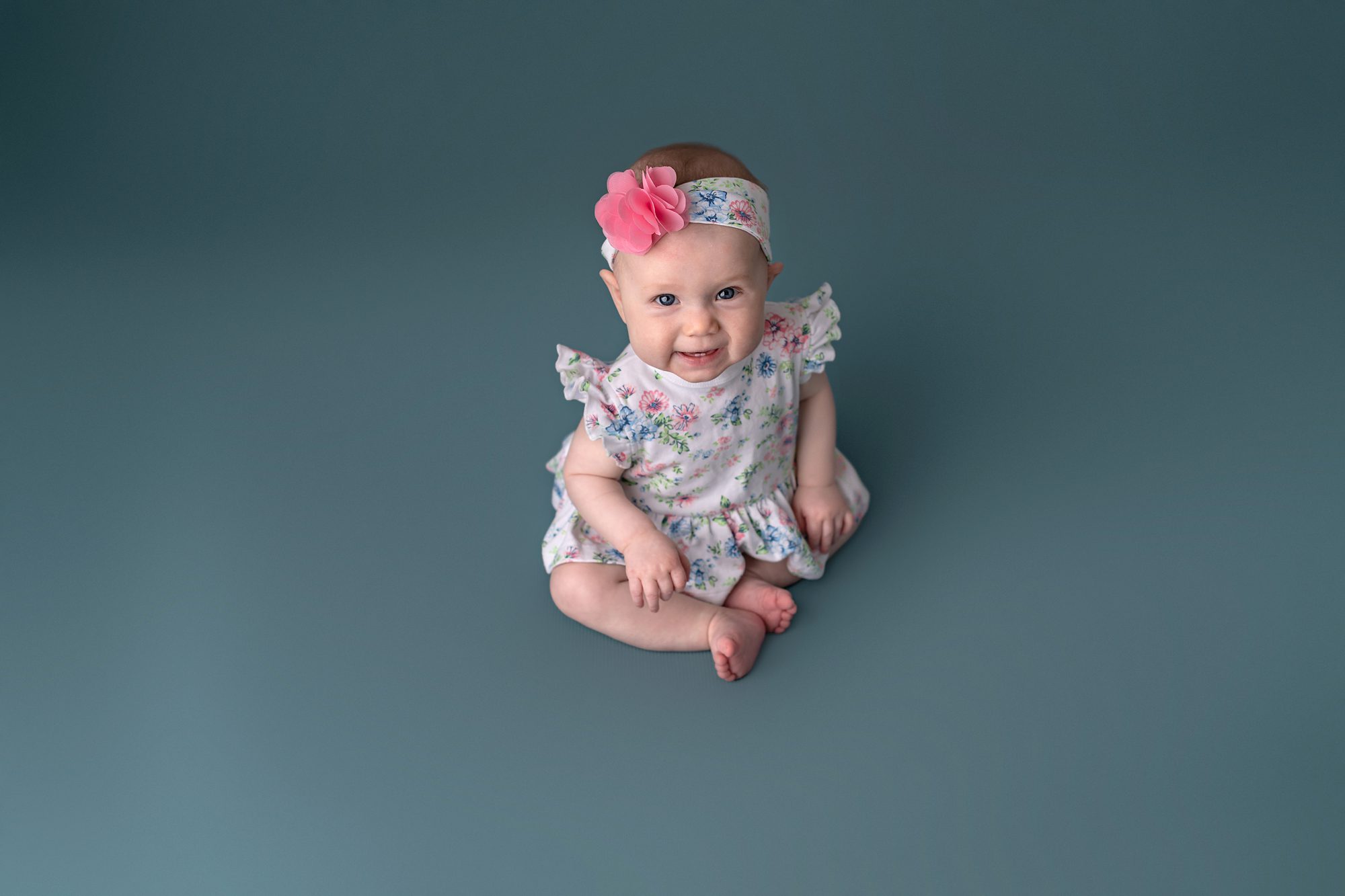 Baby girl sitting and smiling on teal background, photographed at Kansas City photography studio Faces You Love
