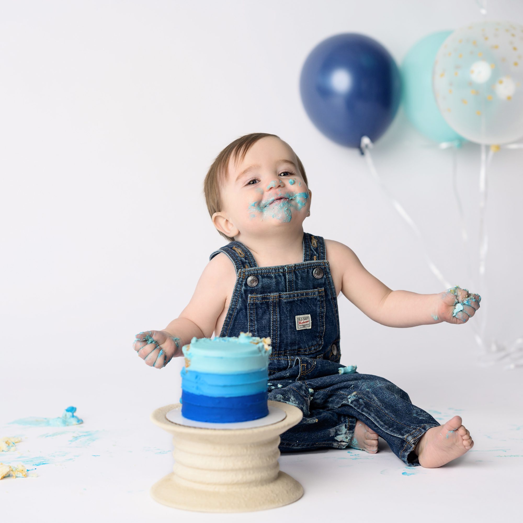Baby boy cake smash session at the Faces You Love Photography Overland Park studio. Cake is shades of blue, with balloons in the background to match. Baby has blue frosting on his face.