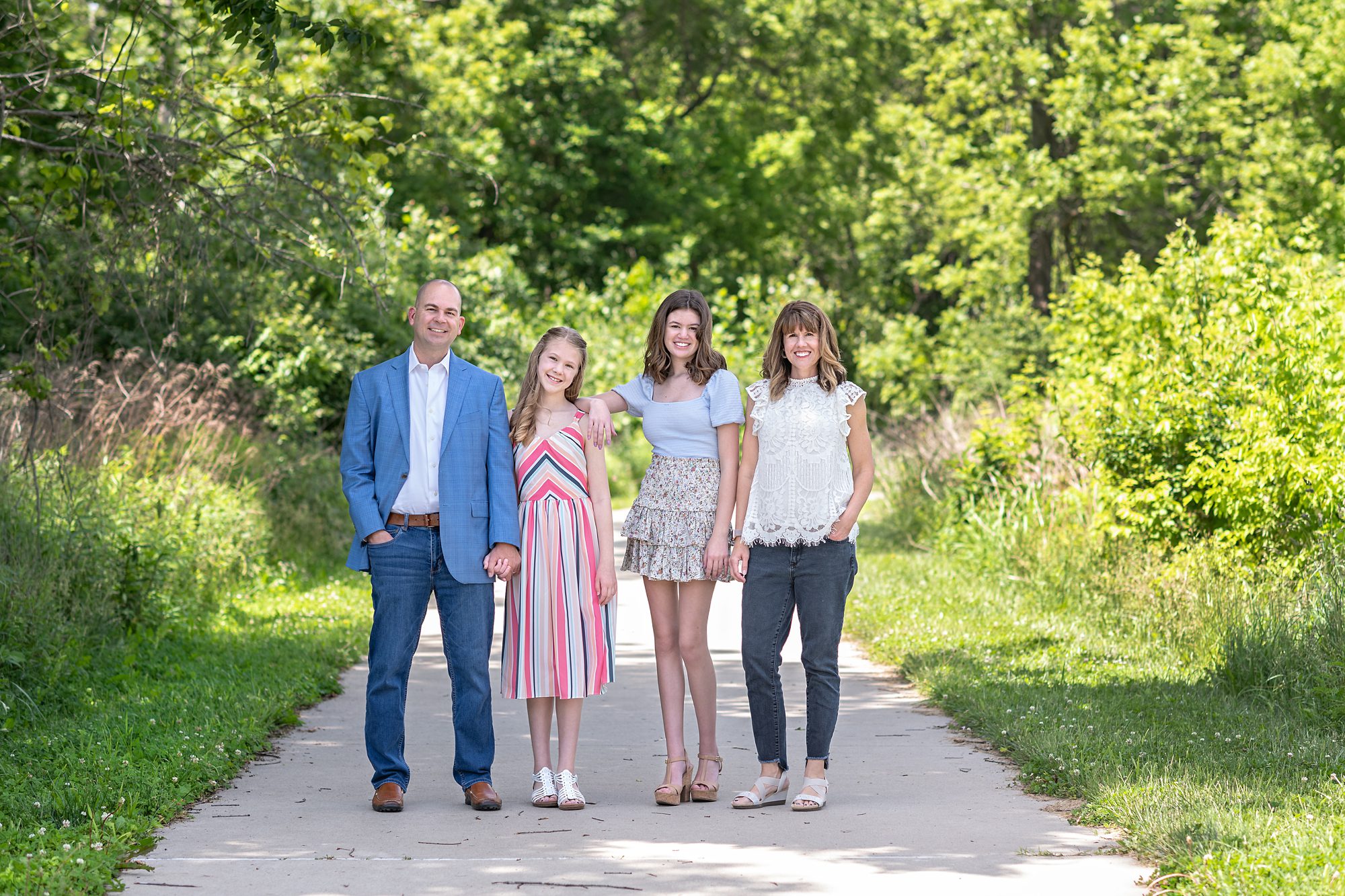 Family standing on a path, smiling for a family portrait. Mom and dad on the ends and two sisters in the middle. Photographed by Helen Ransom of Faces You Love in Kansas City.