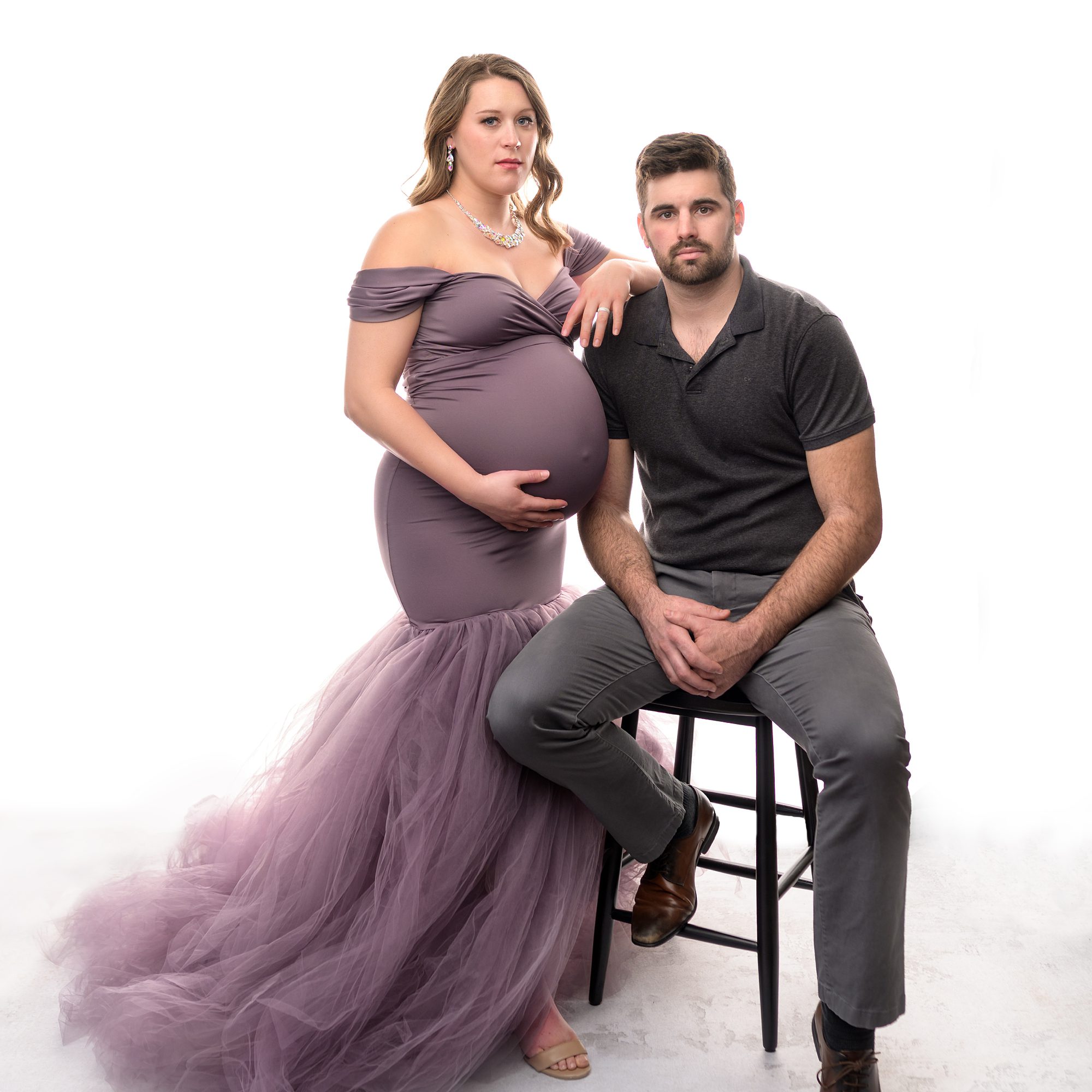 Couple maternity pose, photographed at Faces You Love in Overland Park. Pregnant mom is in a purple, mermaid style gown and is standing next to her seated husband who is dressed in dark gray.
