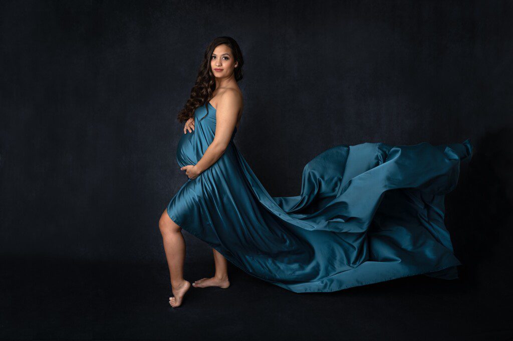 Stunning maternity portrait of mom on a dark background, with teal silk draped on her. Photographed at the Faces You Love Photography studio in Kansas City.