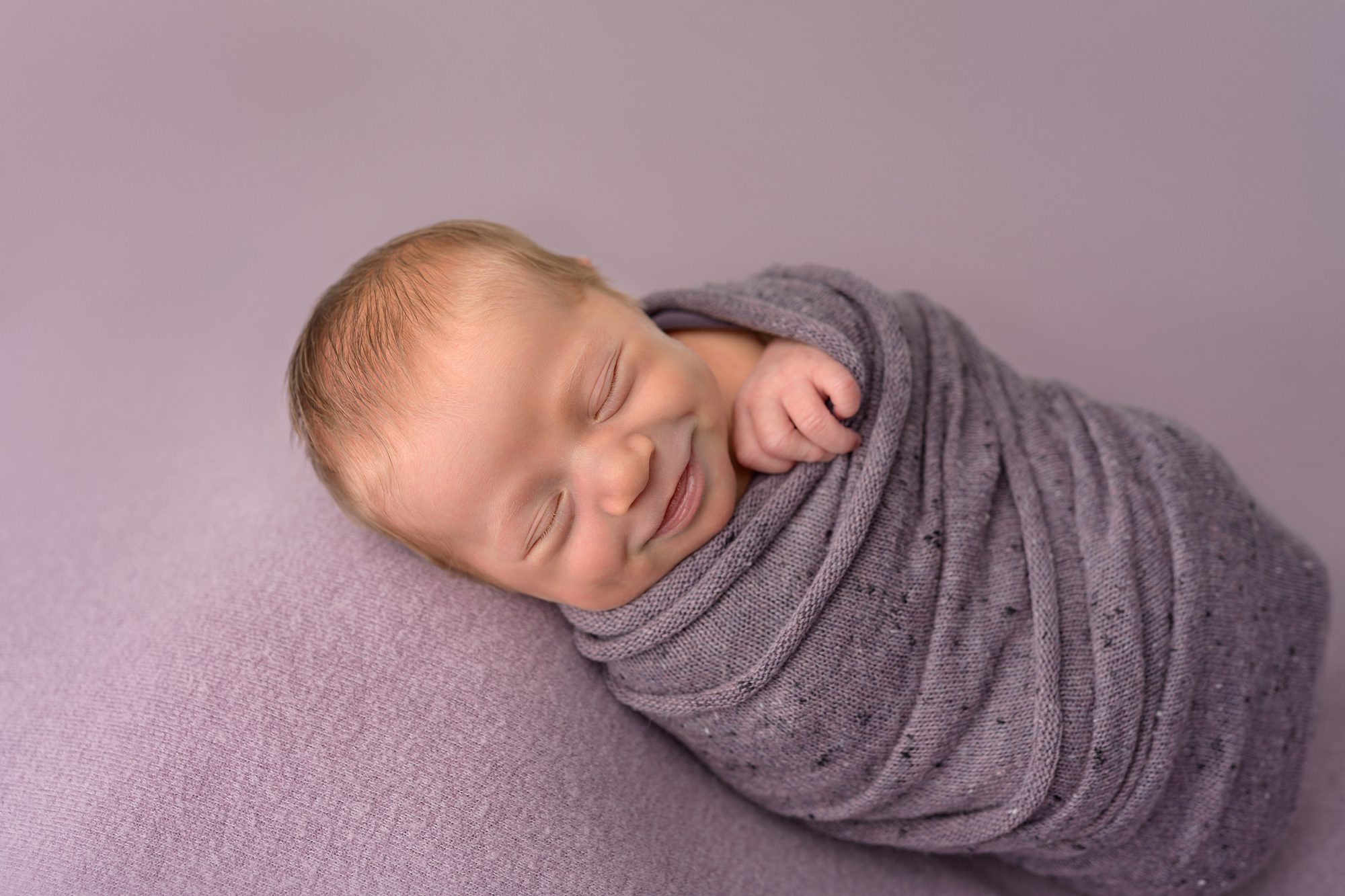 Smiling newborn girl on shades of purple, photographed at the Faces You Love Overland Park portrait studio.