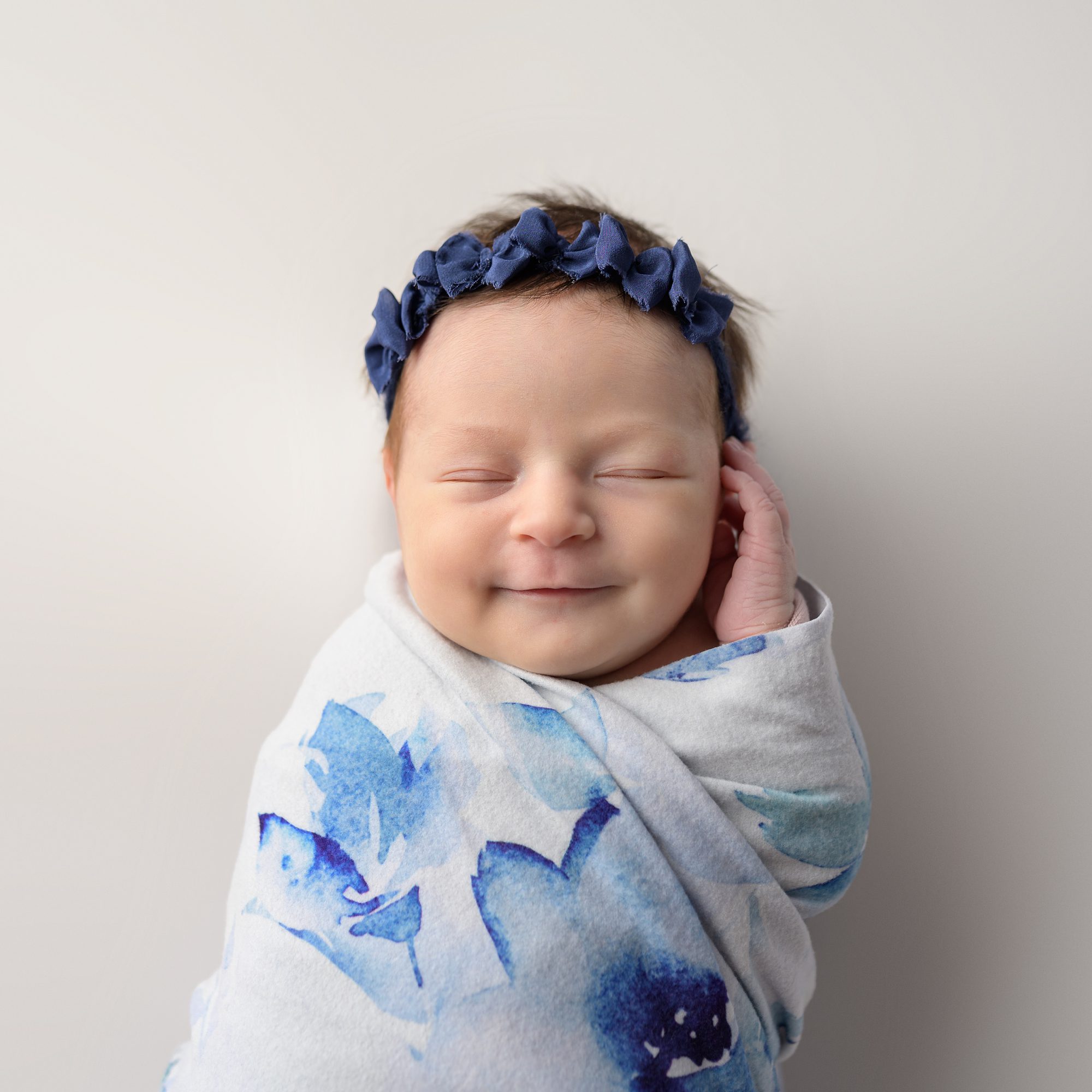 Smiling newborn baby girl, wearing a blue headband and wrapped in a light blue floral swaddle. Photographed by Helen Ransom of Faces You Love in Overland Park.