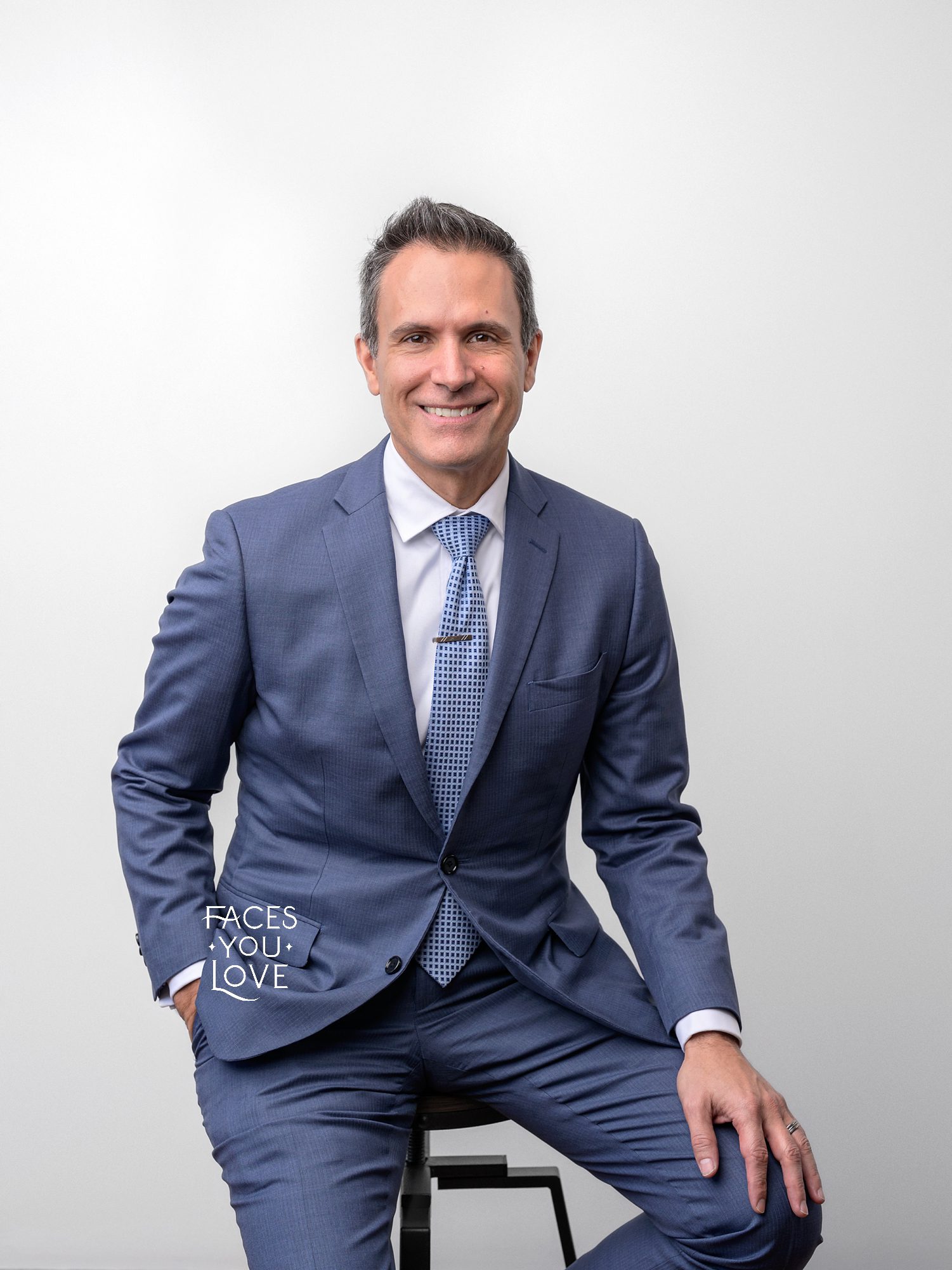Professional male, sitting on a stool in a relaxed pose. He's wearing a blue suit and tie, against a white backdrop.