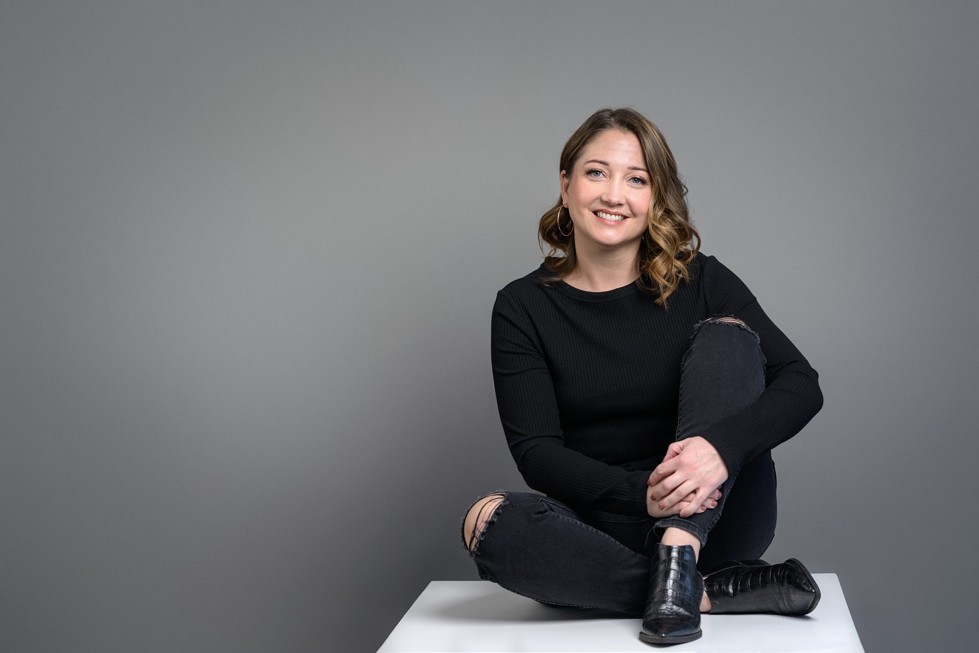 Studio branding photo of smiling woman, dressed all in black, sitting on a white box in front of a gray background. Picture taken in Overland Park, KS at the Faces You Love Photography studio.