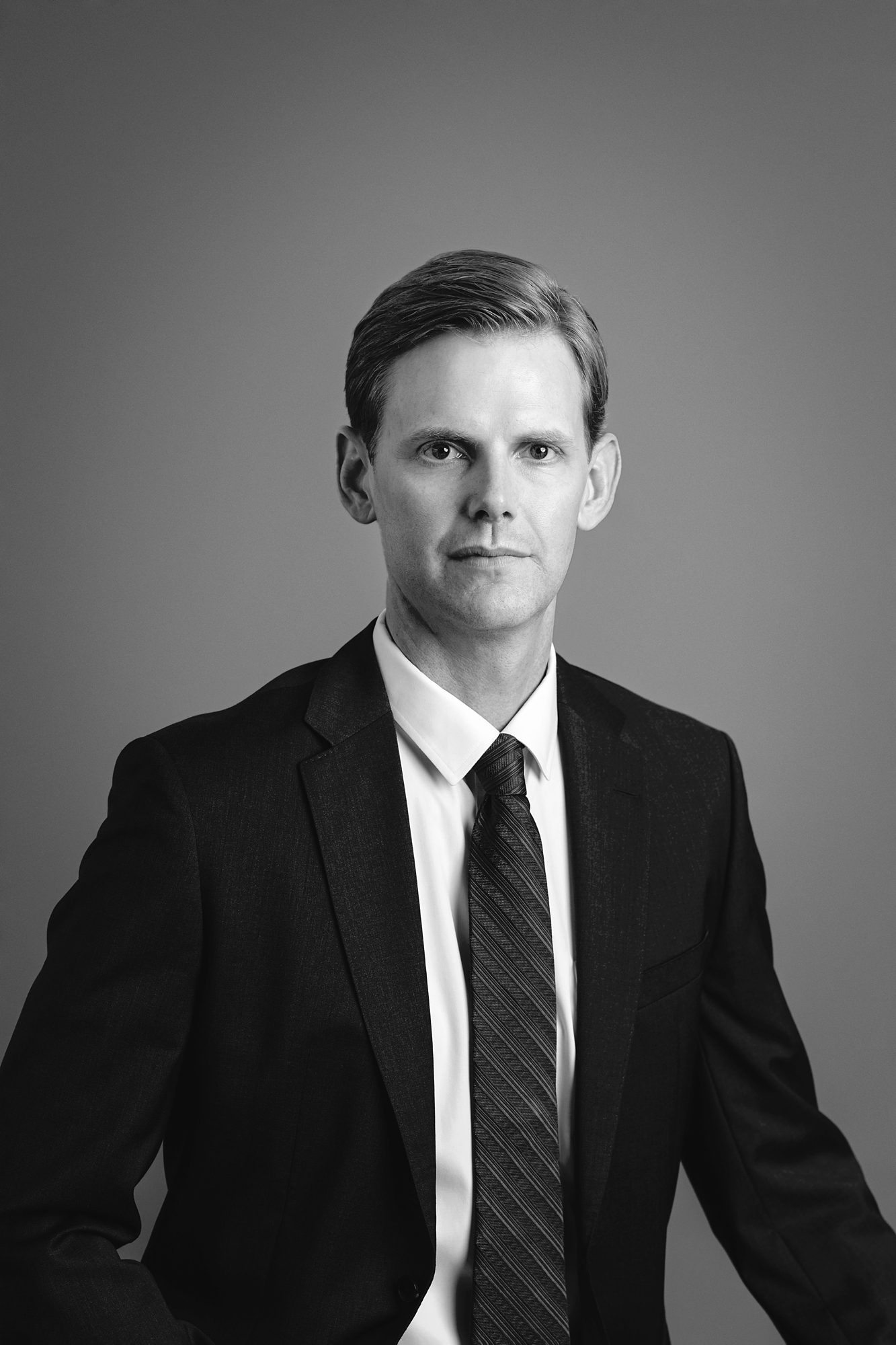 Black and white studio headshot portrait of a man. He has a serious expression on his face, and is wearing a dark colored suit and tie with a white shirt. Photographed by Overland Park photographer Faces You Love.