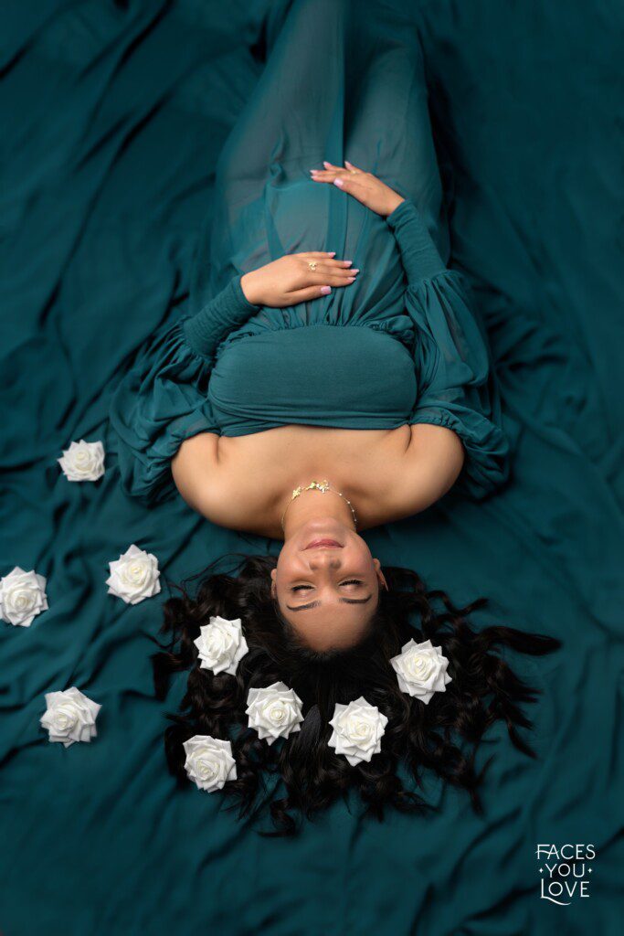 Overhead female maternity photograph, captured on a green background. She's wearing a flowy, green gown with white flowers in her hair. Photographed at Faces You Love studio in Overland Park.